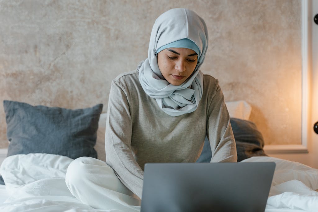 A woman wearing a hijab sits on a bed, concentrating on her laptop. Ideal for home study or remote work themes.