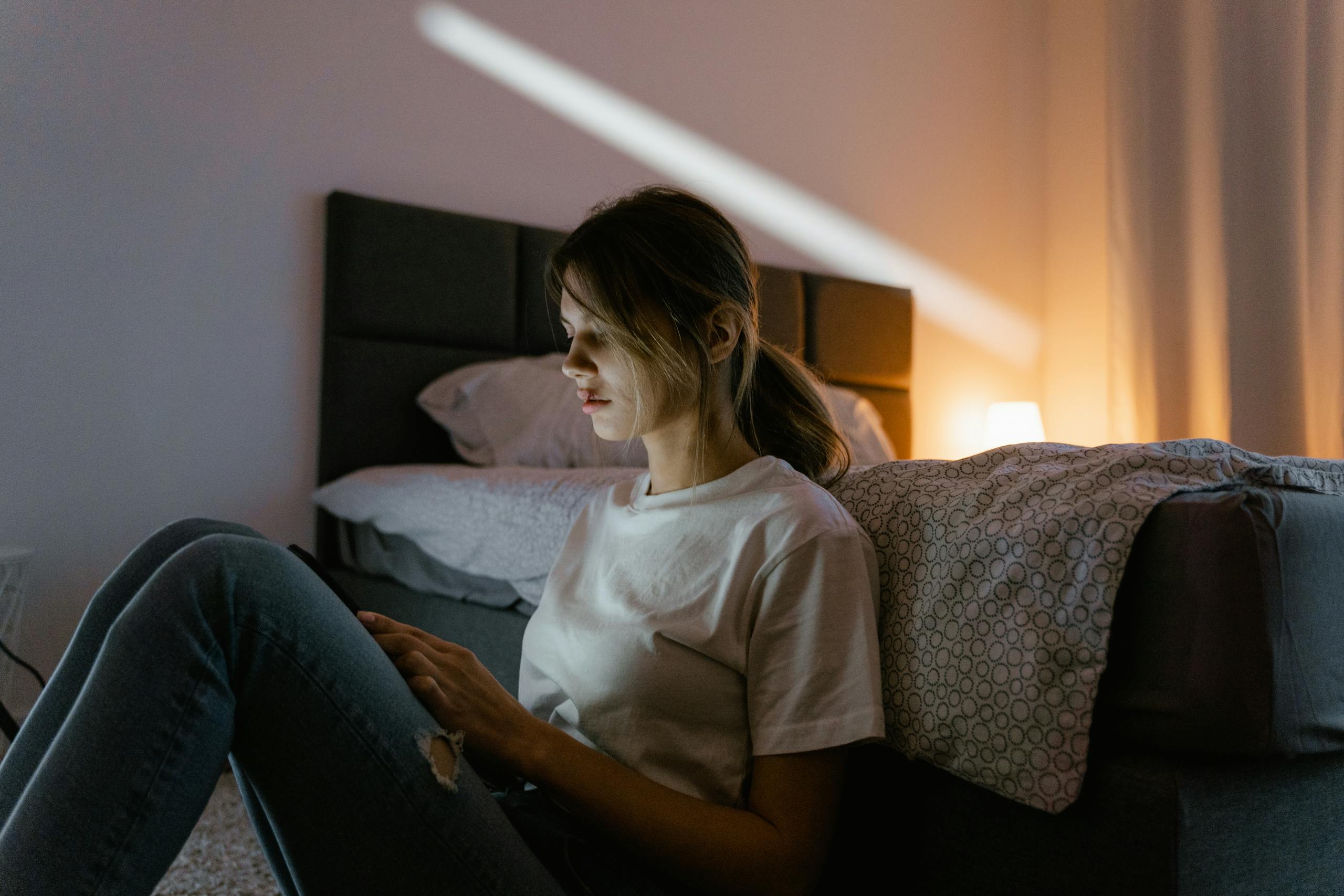 A young woman in a white shirt sits against a bed using a smartphone in a dimly lit bedroom.